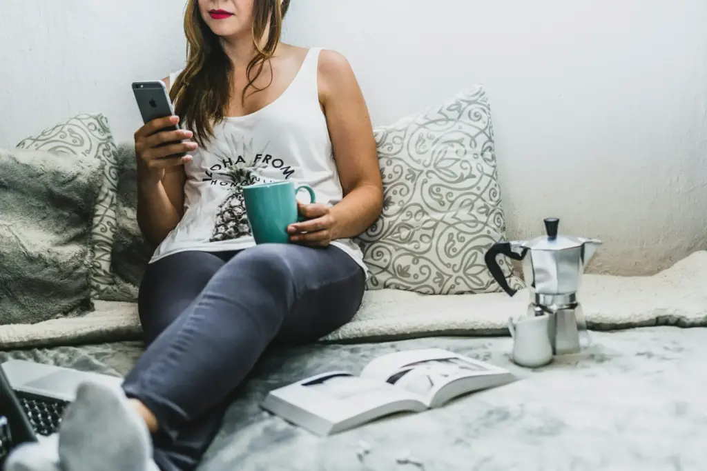 Woman reading her iPhone in one hand with a mug in the other. An open book, Macbook, moka pot, and small creamer pitcher are laid out beside her while she leans against pillows.